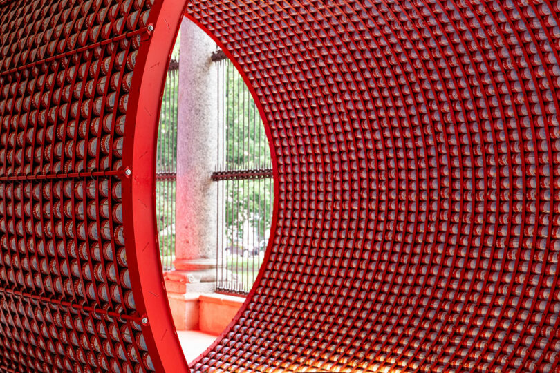 Close-up view of a large cylindrical red metal structure with a grid pattern, looking out onto stone columns and greenery through iron bars.