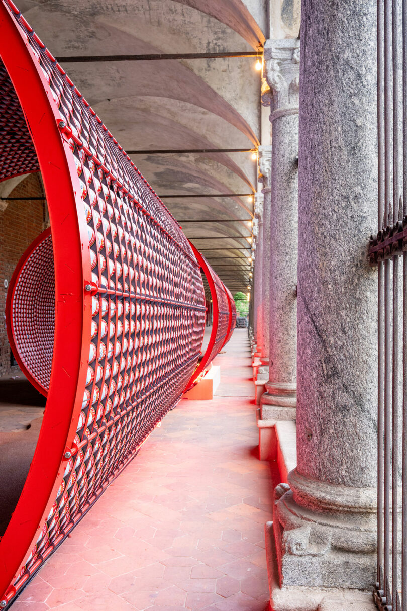 A large red cylindrical art installation with a patterned surface is displayed under an arched stone hallway with columns.