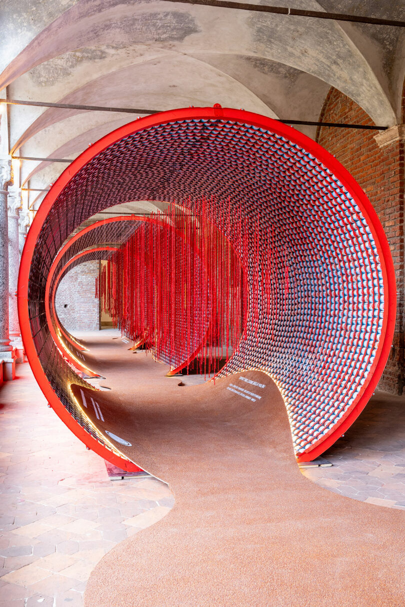 A large red circular installation with a mirrored interior and hanging red strings is displayed in an arched hallway with brick walls and tiled flooring.