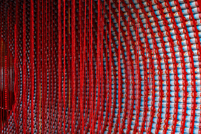 Rows of red cords hang vertically in front of a wall stacked with aluminum cans arranged in neat, curved rows.
