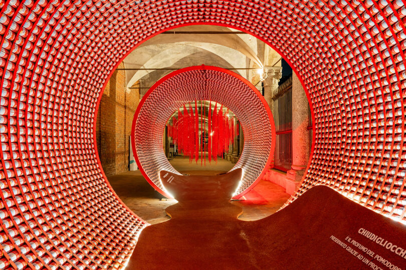 A brightly lit installation features concentric circular structures lined with red cups, creating a tunnel effect inside an industrial space with brick walls and arched ceilings.