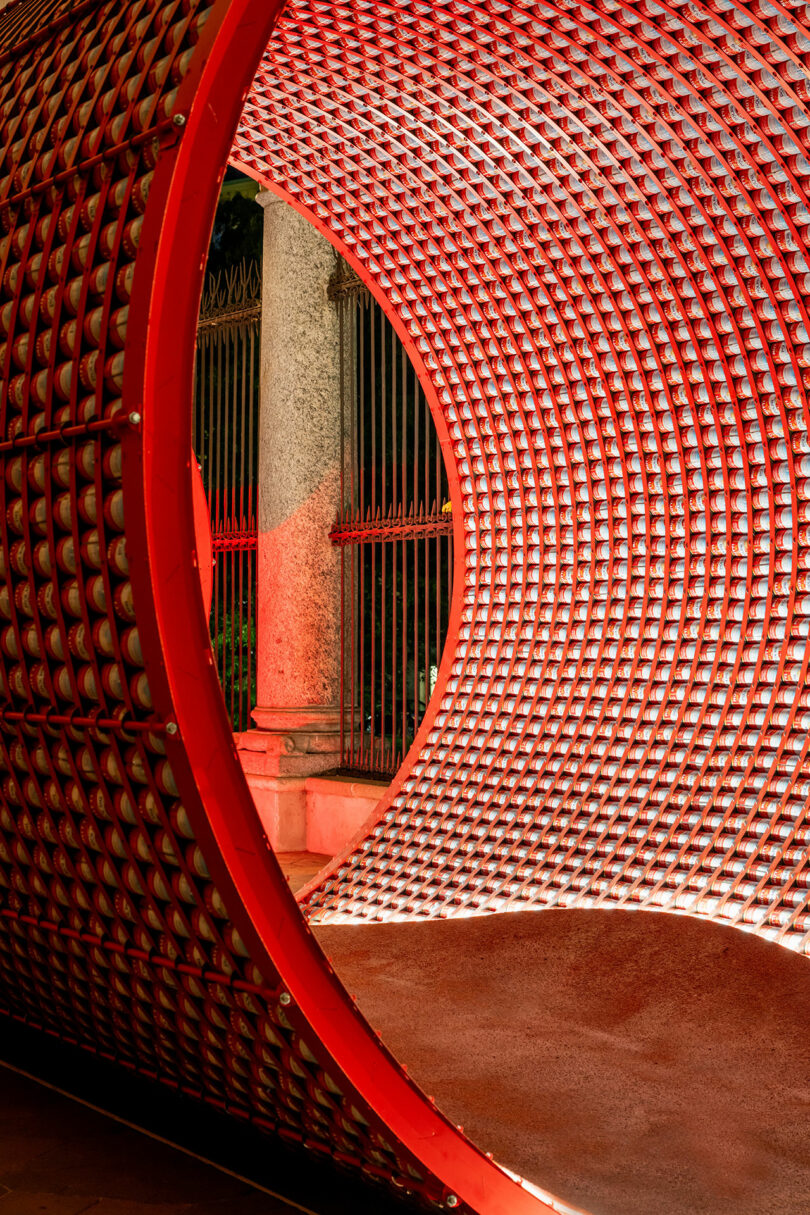 A large, cylindrical tunnel structure with a red grid pattern, illuminated by light, stands next to a metal fence and stone columns.