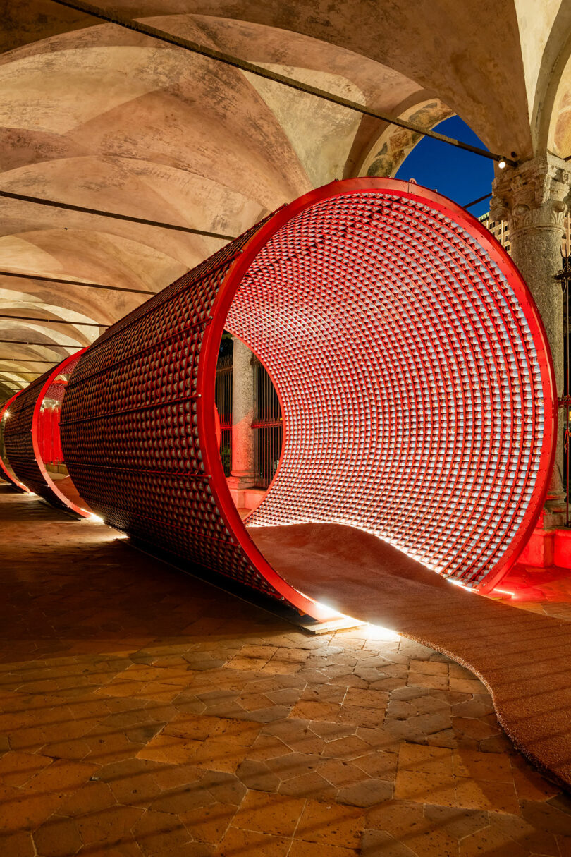 A large, red cylindrical tunnel structure with a patterned grid surface is displayed indoors under arched ceilings, illuminated by lights on the floor.