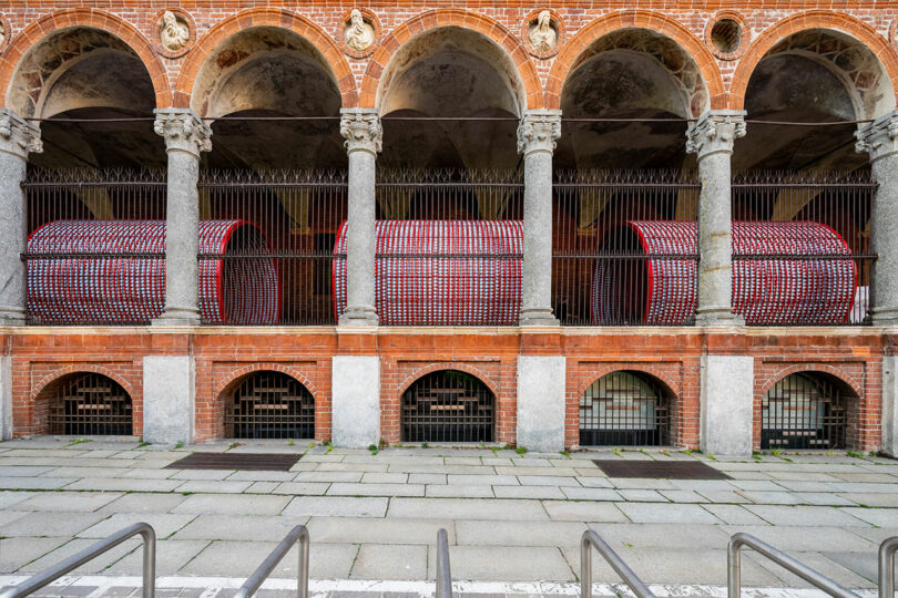 A brick and stone building with arches and columns displays three large cylindrical art installations covered in red and white patterns behind metal bars.