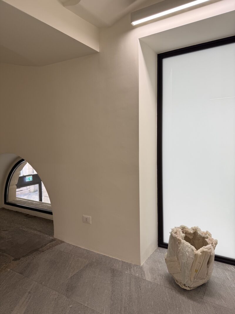 A minimalist room with light walls, a large frosted window, a small arched window, stone flooring, and a textured, cylindrical sculpture in the corner.