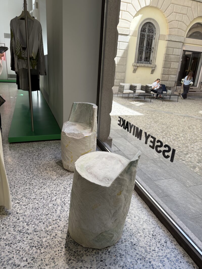 Two sculptural, abstract chairs made of a white, textured material are positioned near a large window in a modern store, with a street scene visible outside.