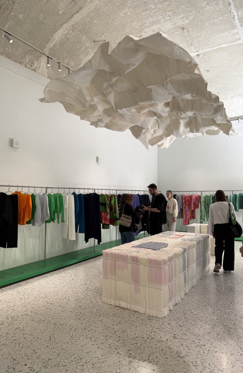 Clothing store interior with colorful garments on racks, a textured ceiling installation, and people browsing near a fabric-covered display table.
