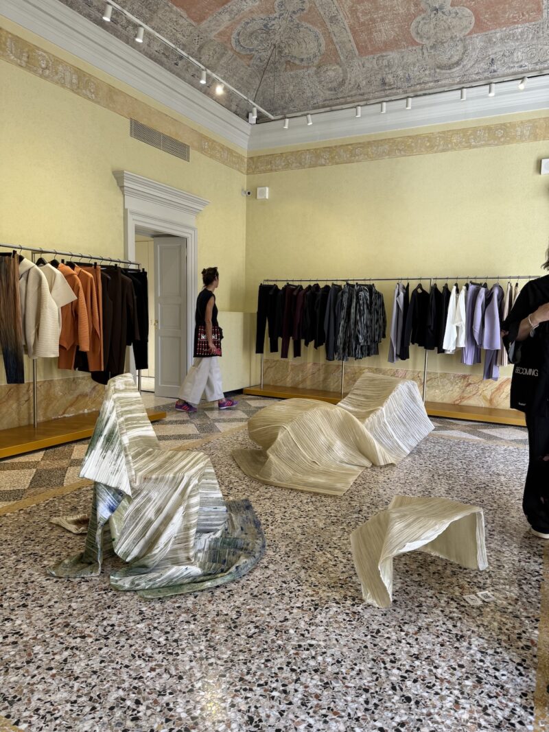 A person walks through a clothing showroom with racks of jackets and shirts along the walls and sculptural, folded bench-like pieces displayed on the floor.