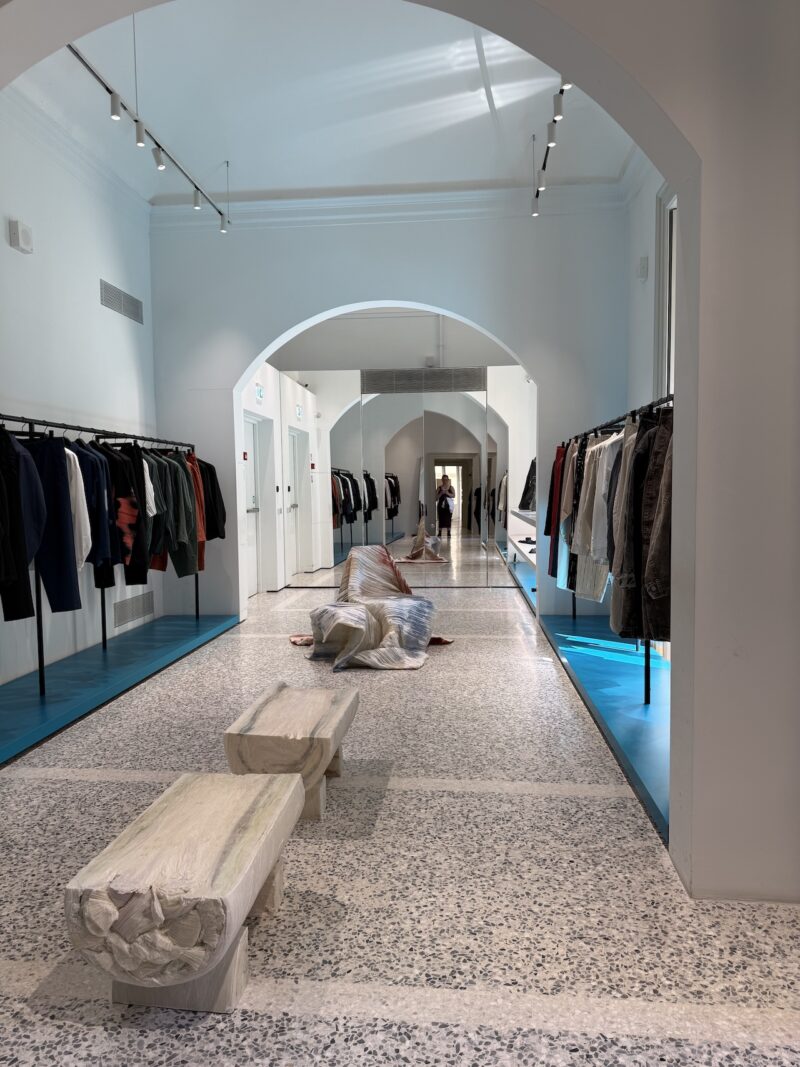 Clothing store interior with racks of clothes on both sides, a terrazzo floor, two benches, and a person lying on the floor in the center hallway.