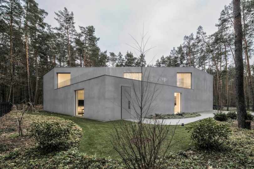 Modern corner gray concrete house by KWK Promes with large windows, surrounded by grass, bushes and tall pines in a wooded area.