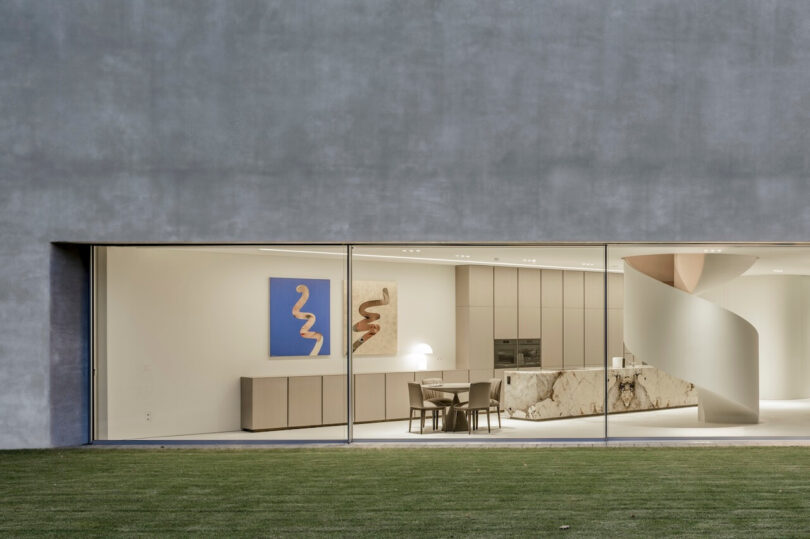 A modern kitchen and dining room designed by KWK Promes features minimalist decor, marble countertops, a spiral staircase and large floor-to-ceiling windows overlooking a lush green lawn.