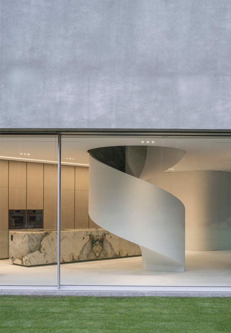 A modern interior by KWK Promes with a large glass window, a white spiral staircase, light wood cabinetry and a marble kitchen island. The green grass is visible outside.