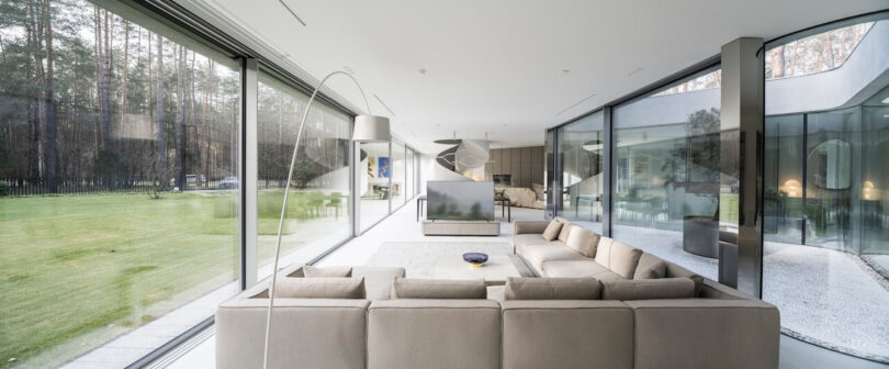 Modern living room designed by KWK Promes, with a large beige sofa, glass walls and a view of a green lawn and forest. Minimalist decor and plenty of natural light fill the space.