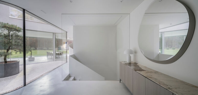 Minimalist modern interior by KWK Promes with large glass windows, marble console, round mirror and white lamp, with outdoor garden visible through elegant windows.