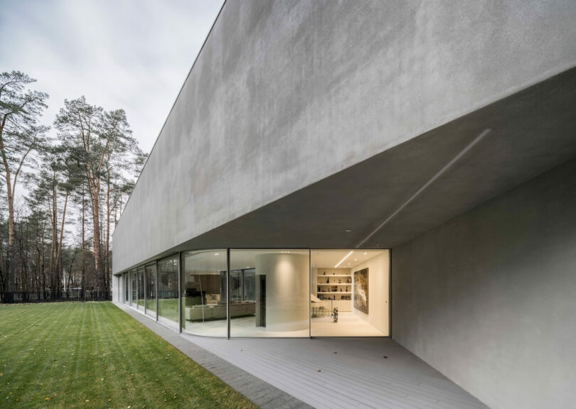 Modern minimalist building by KWK Promes with large glass windows and concrete exterior, next to a beautifully trimmed lawn and bordered by a wooded area.
