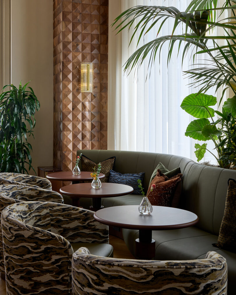 A modern lounge area by LEGEARD STUDIO features round tables, patterned armchairs, a curved green sofa, small vases with flowers, and large leafy plants by tall windows with sheer curtains.