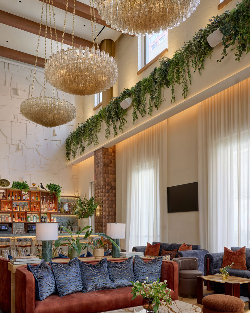 Modern hotel lobby by LEGEARD STUDIO featuring high ceilings, large chandeliers, hanging greenery, plush seating, a stylish bar area, and tall windows draped with sheer curtains.