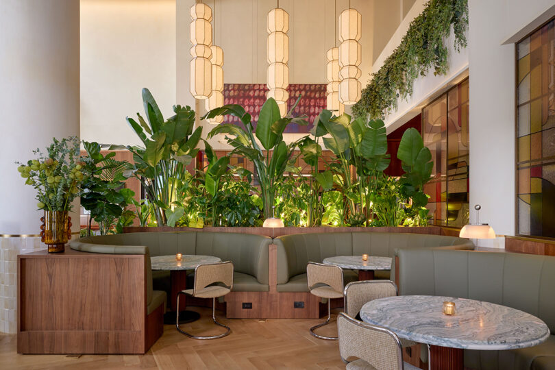 A modern restaurant interior by LEGEARD STUDIO features curved green upholstered booths, marble tables, wicker chairs, potted plants, hanging lights, and leafy greenery as a stylish divider.