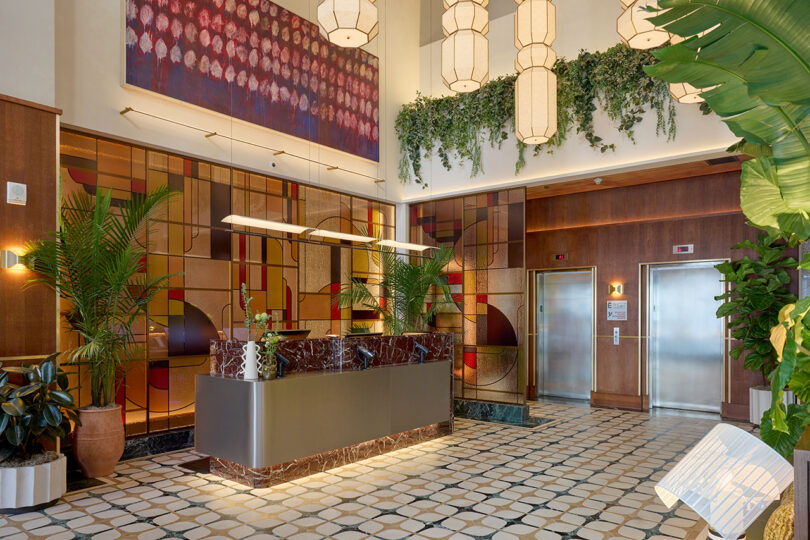 Hotel lobby with a marble reception desk by LEGEARD STUDIO, two elevators, potted plants, geometric wall art, hanging lights, and decorative foliage.