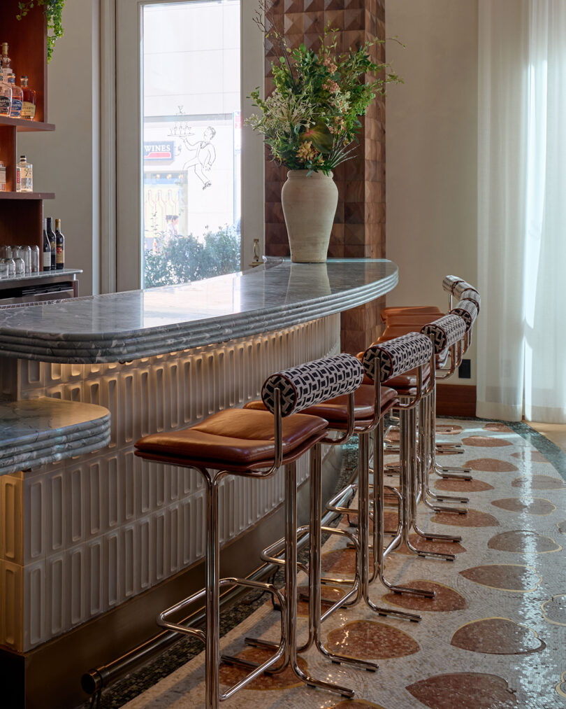 A modern bar area by LEGEARD STUDIO features four leather and chrome barstools, a marble countertop, decorative tile flooring, and a large vase of flowers on the counter.