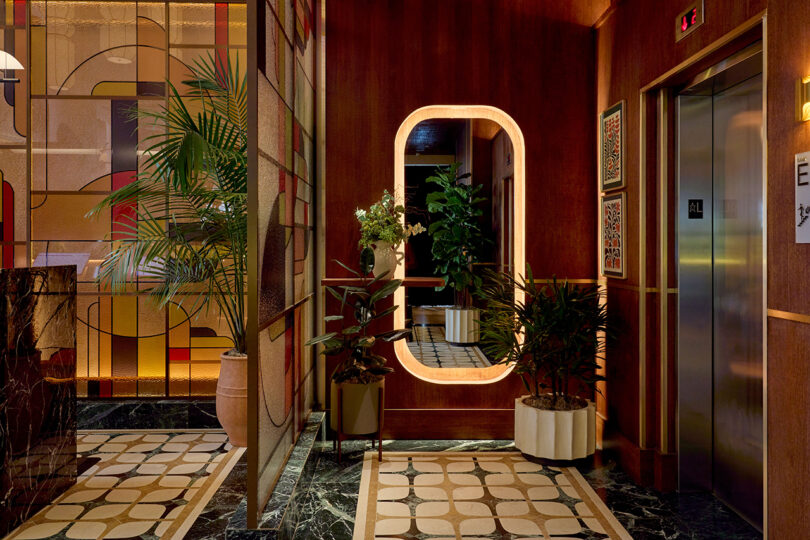 A hallway by LEGEARD STUDIO features patterned tile flooring, potted plants, a large illuminated mirror, an elevator, and decorative wall panels with striking geometric designs.