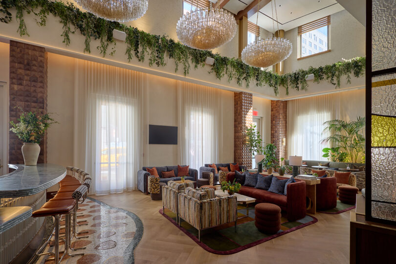 Modern lounge area by LEGEARD STUDIO with large windows, velvet sofas, bar stools, hanging chandeliers, and greenery along the ceiling. Natural light fills the room, creating a bright and inviting space.