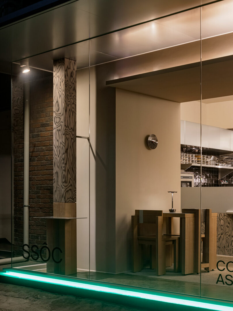 Modern restaurant interior viewed through large glass windows, featuring wooden tables and chairs, exposed brick, and illuminated green strip lighting along the floor.