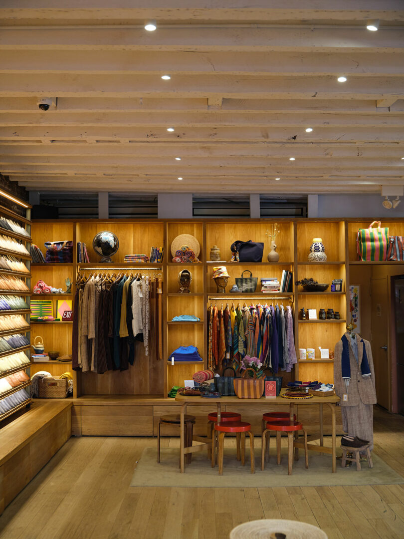 A neatly organized clothing store displays folded shirts, hanging garments, accessories, and decor items on wooden shelves with a central table and red stools.