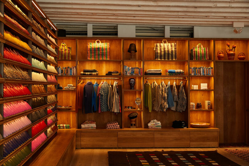 A neatly organized boutique displays colorful folded textiles on shelves, clothing hanging on racks, and various accessories arranged on wooden cubby shelves.