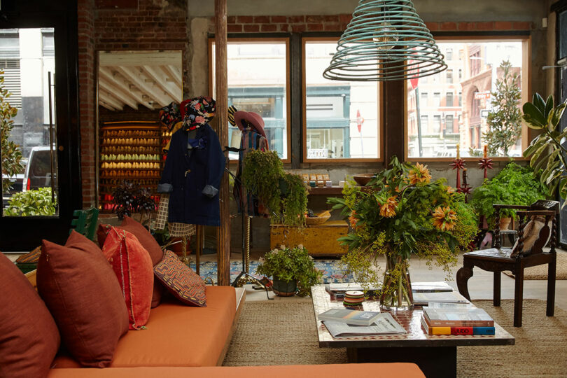 A cozy, eclectic lounge with orange sofas, vibrant cushions, a coffee table with books and flowers, leafy plants, and a clothing display near large windows letting in natural light.