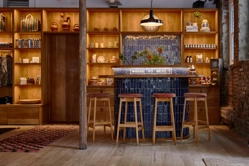 A cozy bar area with four wooden stools, a tiled counter, shelves displaying various items, and a "WHERE TO NEXT?" sign on the counter.