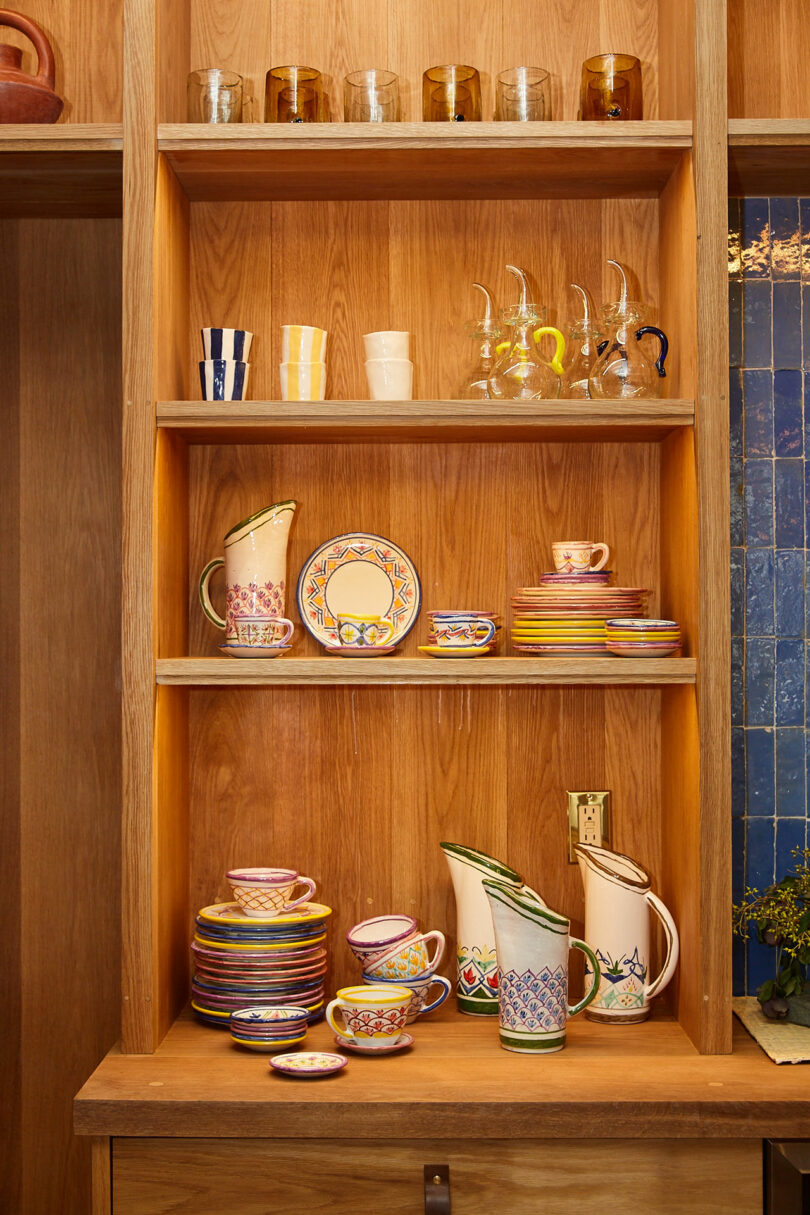 Wooden shelves displaying glassware, cups, colorful ceramic plates, mugs, and pitchers with intricate patterns and designs.