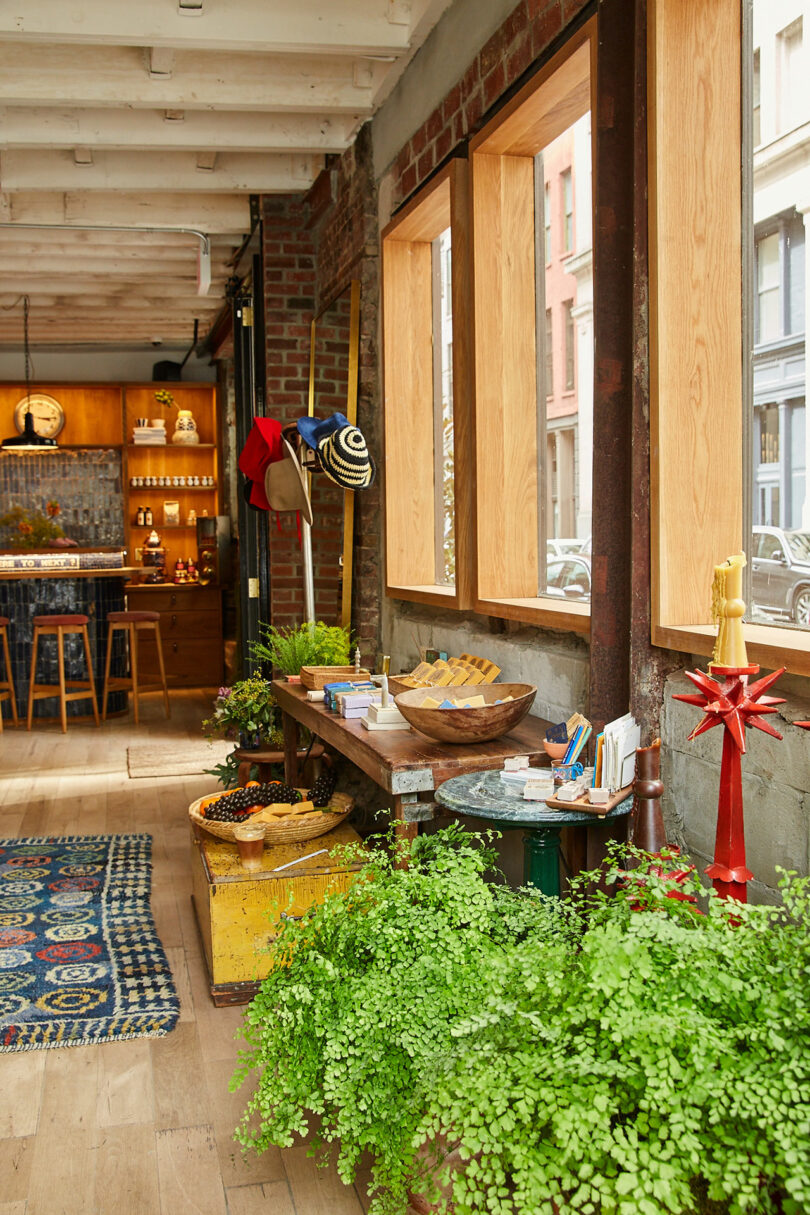 A rustic shop interior with wooden floors, potted plants, a wooden table displaying bowls and goods, hats on the wall, and shelves with jars and bottles in the background.