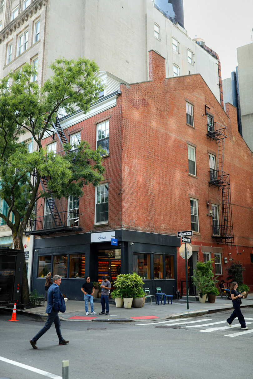 A red brick corner building with a ground floor café, fire escapes, and people walking on the sidewalk at an urban intersection.
