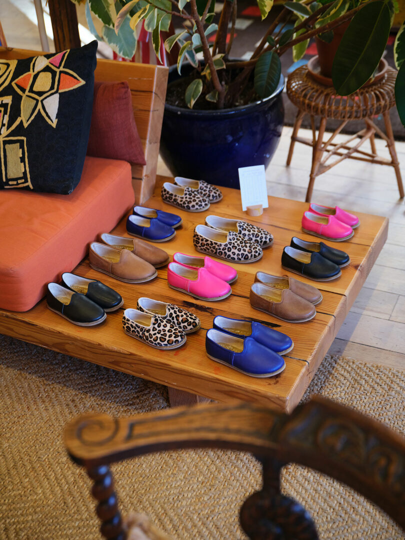 A variety of children’s slip-on shoes in different colors and patterns, including pink, blue, leopard print, black, and brown, are displayed on a wooden platform indoors.