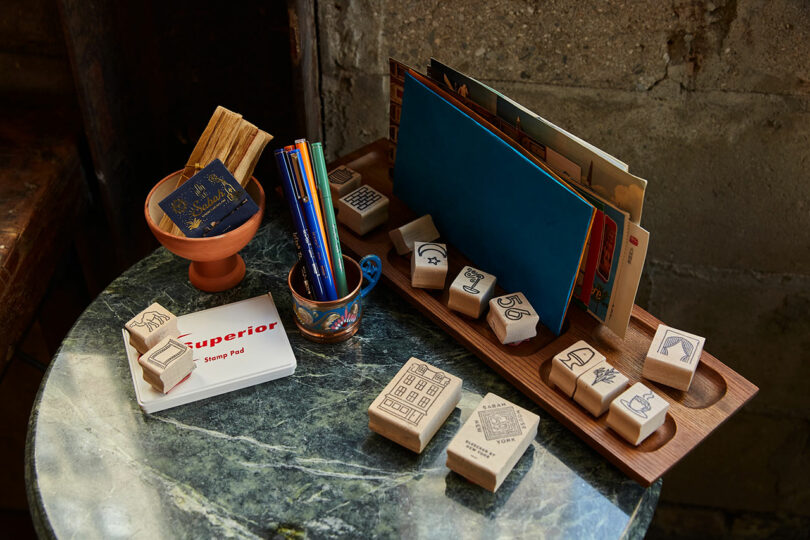 A round marble table displays wooden stamps, colored pencils in a cup, a notepad, books, and postcards against a stone wall.
