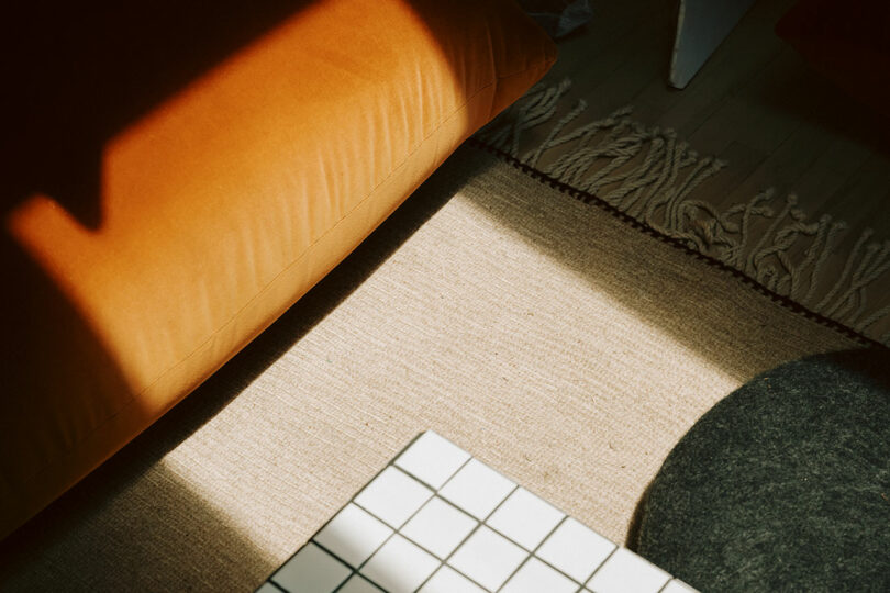 Sunlight casts shadows on a beige rug next to an orange cushion, a gray round object, a white checkered surface, and a fringed edge of another rug.