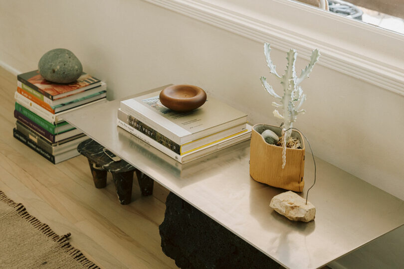 A metal bench holds stacked books, a wooden bowl, and a potted succulent. More books and a stone rest on the floor beside the bench, near a window.