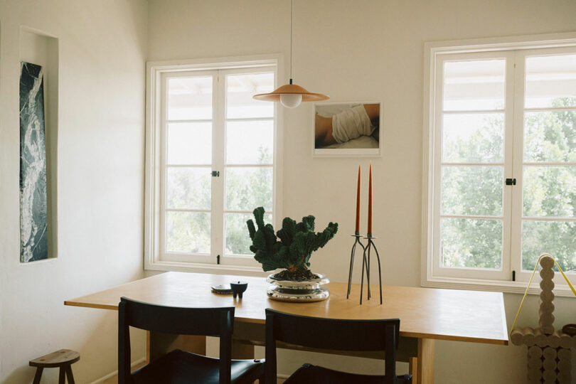 A minimalist dining area with a wooden table, black chairs, two tall candles, a potted plant, and two large windows letting in natural light.