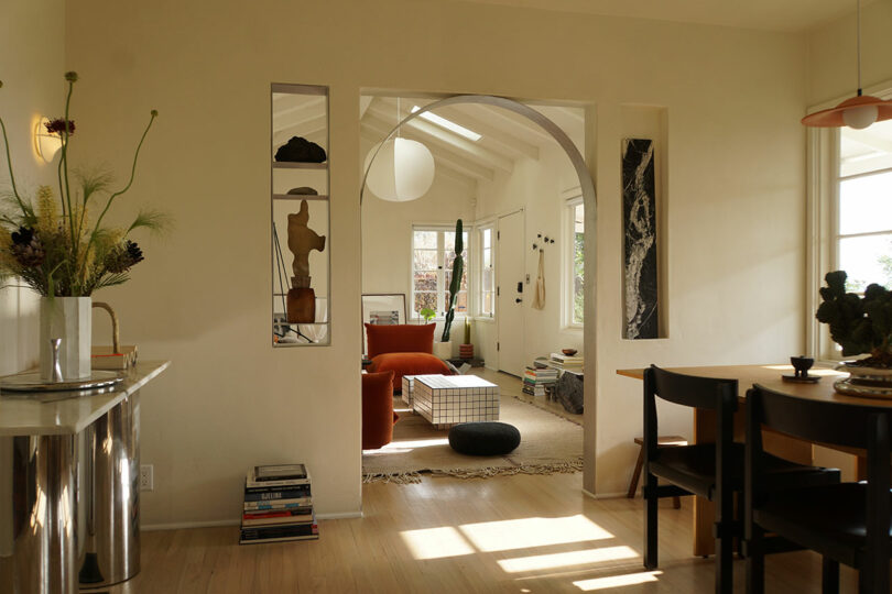 A sunlit living and dining area with neutral walls, modern furniture, a round archway, books, and decorative items on shelves and tables.