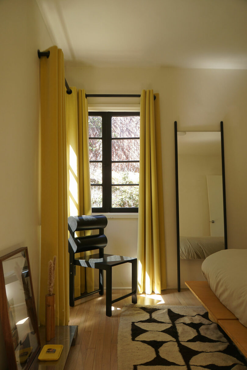 Bedroom with yellow curtains, a window, a black chair, a tall mirror, a geometric rug, a low bed, and framed artwork on the floor. Sunlight streams through the window.