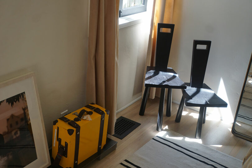 Two black chairs sit in a sunlit corner near a small window, with a yellow suitcase and a framed picture propped against the wall.