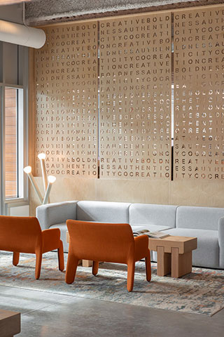 A modern lounge area with orange chairs, a gray sofa, wooden tables, and a large wall art piece featuring a word search design.