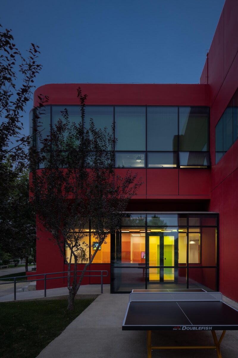 A modern red building with large windows, illuminated interior, and a ping pong table outside on a concrete path at dusk.