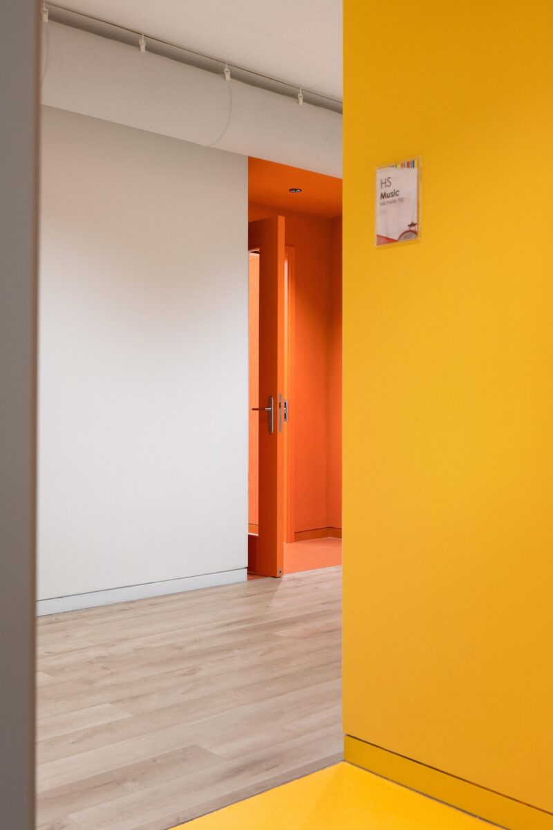 A hallway with yellow walls, light wooden flooring, and an orange door slightly open leading to another room; a sign labeled "HS Music" is mounted on the yellow wall.