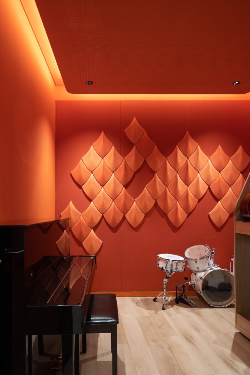 A modern music room with orange acoustic wall panels, a black upright piano with a bench, and a white drum set on a light wood floor.