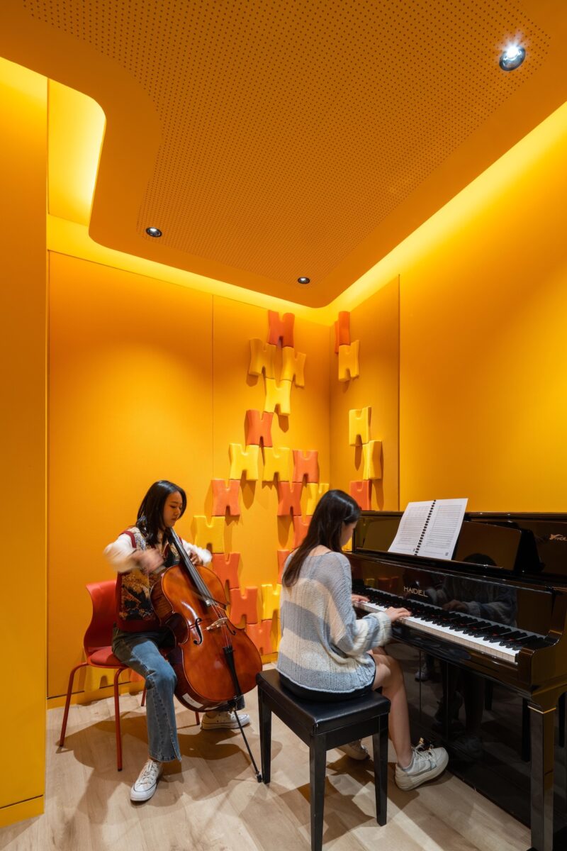 Two young women play music in a bright yellow soundproof room; one plays the cello and the other plays the piano.
