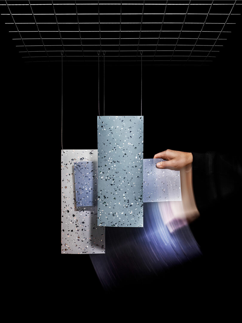 A hand moves behind three suspended rectangular Tarkett terrazzo panels of varying sizes and colors, with a grid structure above against a black background.