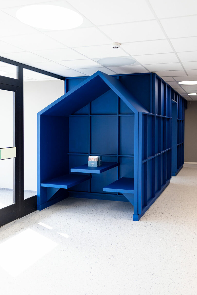 A built-in, house-shaped blue alcove with Tarkett flooring features seating and a small table, standing against a wall in a modern hallway. A single book rests on the table.