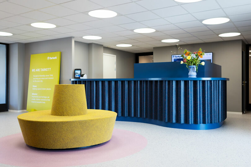 Modern office reception area with a blue desk, digital clock, Tarkett flooring, flower arrangement, and a yellow circular bench on a pink rug. Informational sign is visible on the left wall.
